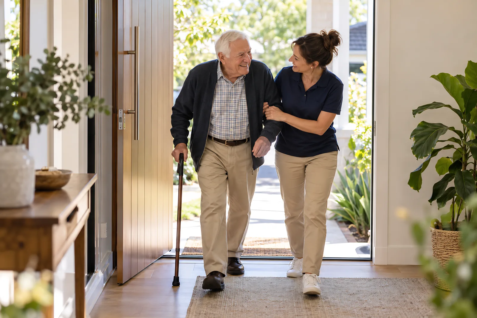 Support worker walking arm in arm with an older man with a walking stick through his front door